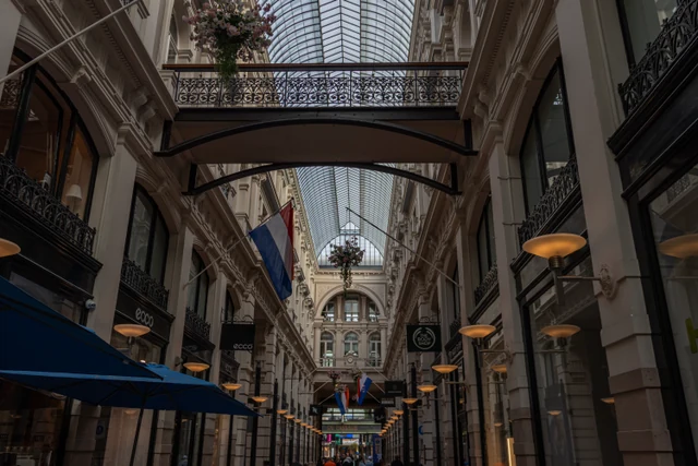 The glass-roofed interior of De Passage in The Hague, with ornate iron balconies, hanging flower baskets, and Dutch flags