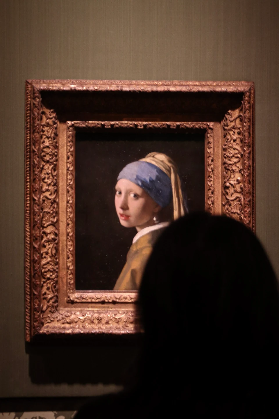 Johannes Vermeer's Girl with a Pearl Earring in its ornate frame, partly obscured by a museum visitor in the foreground