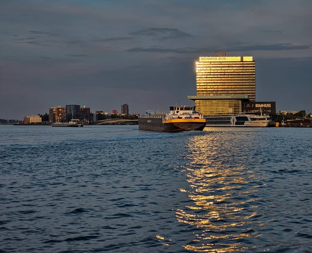 A ferry and riverboats on Amsterdam's waterfront at dusk, with gold hotel lights reflected in the water
