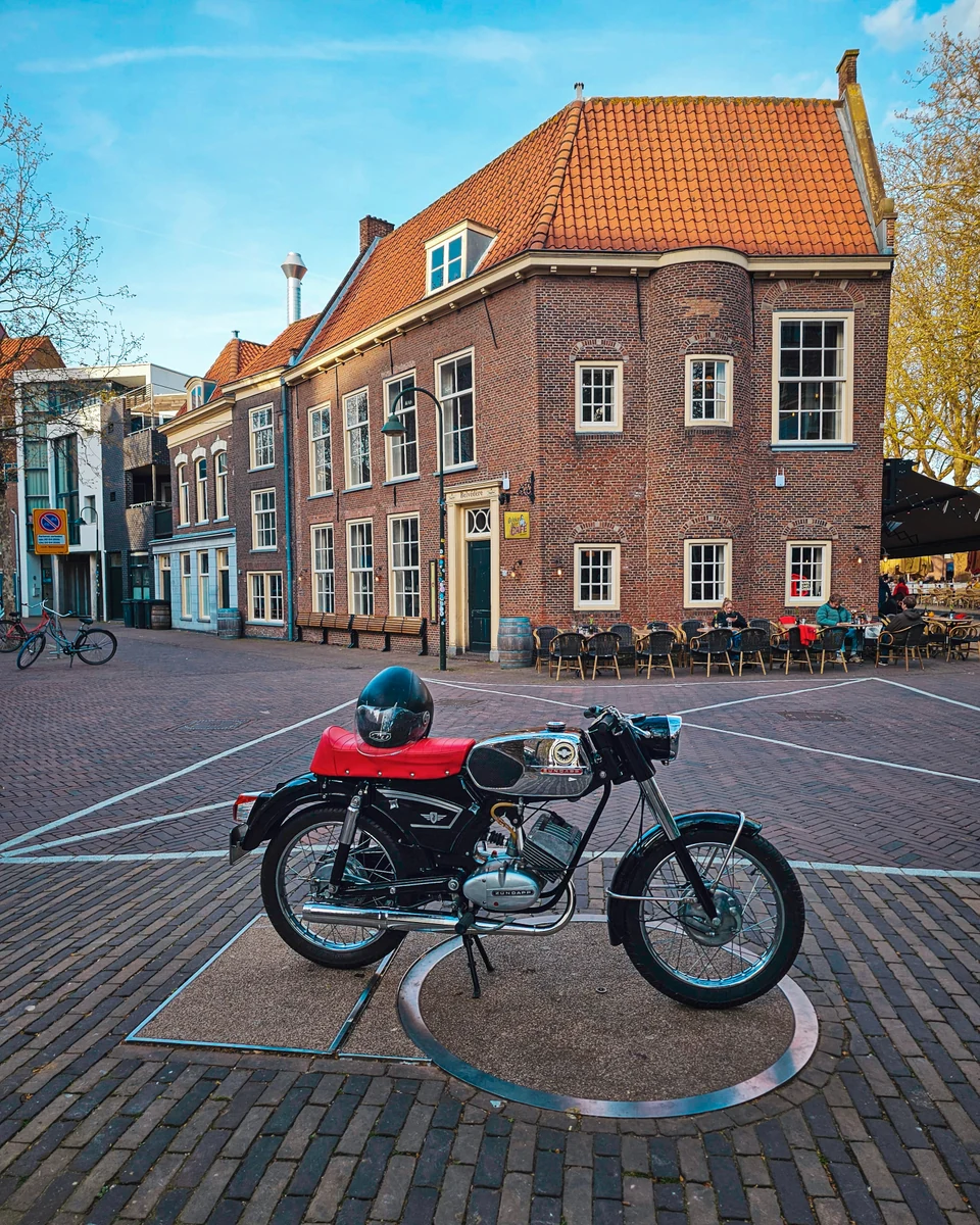 A vintage black Zündapp moped parked in front of a brick building with a tiled roof and outdoor café tables in The Hague