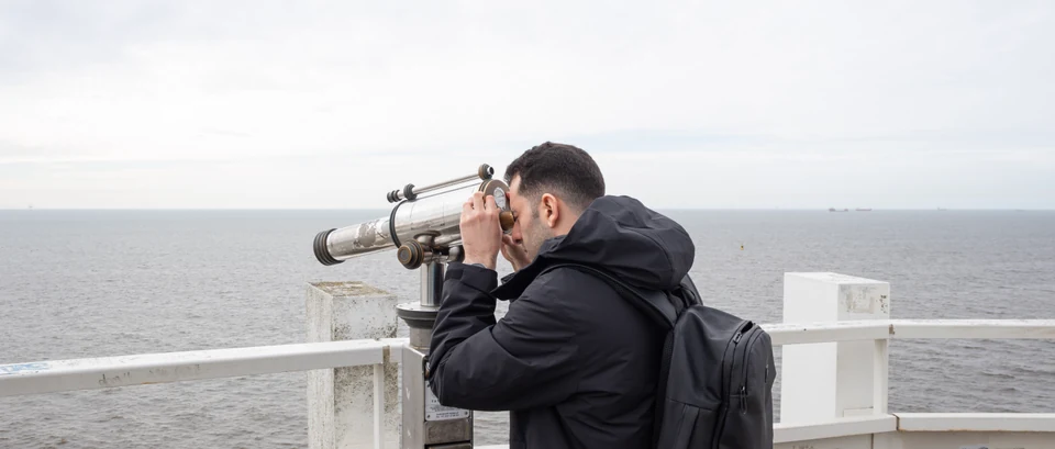 A self-portrait of the author looking through a public telescope across the North Sea, with distant ships on the horizon